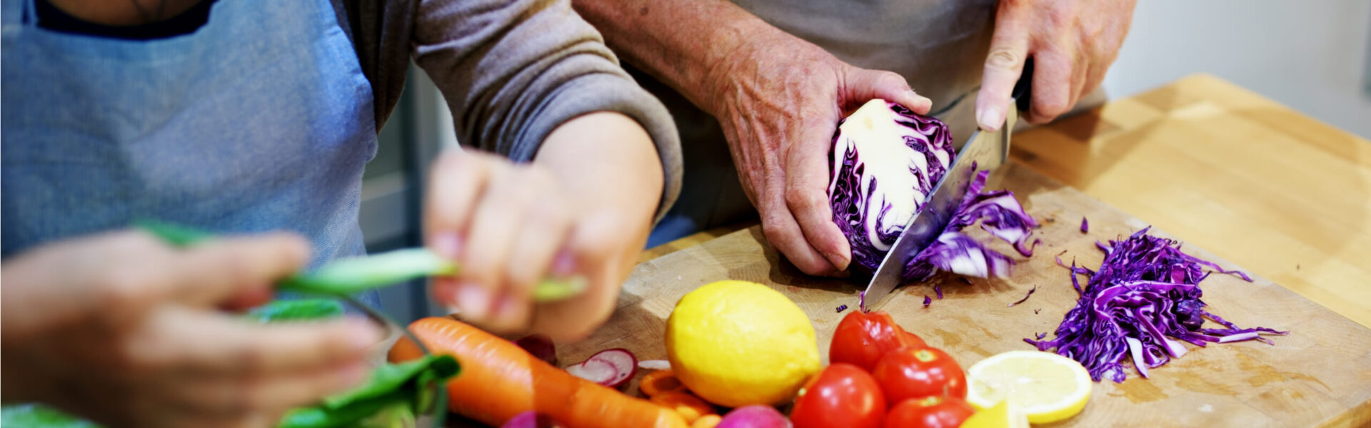 Hands hold fresh vegetables: carrots, onions, tomatoes, beans, corn, and greens, arranged on a dark wooden table.