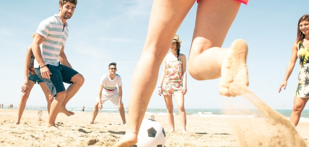 A group of people plays soccer on a sandy beach. One person kicks the soccer ball, while others watch or prepare to participate under a clear blue sky.