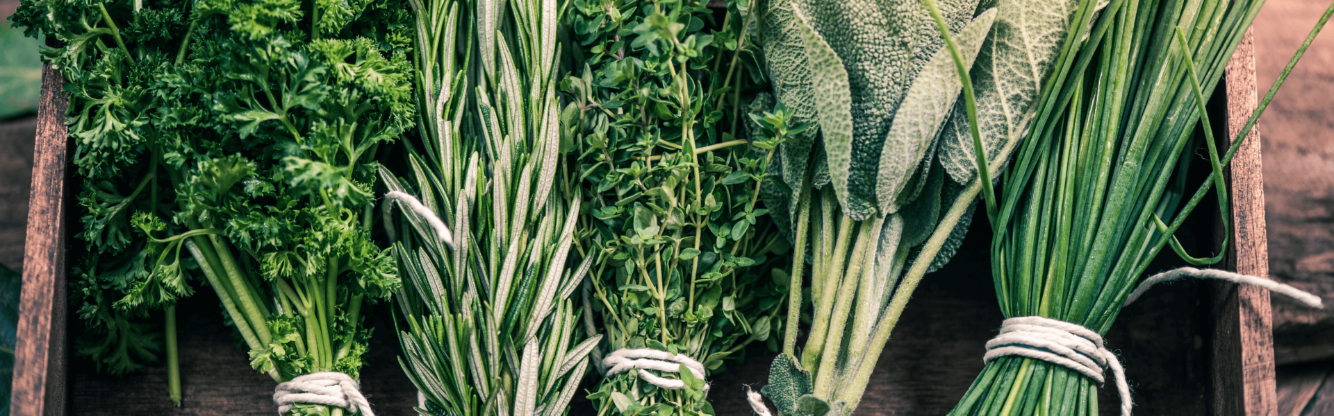 Bundles of fresh herbs, including parsley, rosemary, thyme, sage, and chives, are neatly tied with string and arranged side by side in a wooden box.