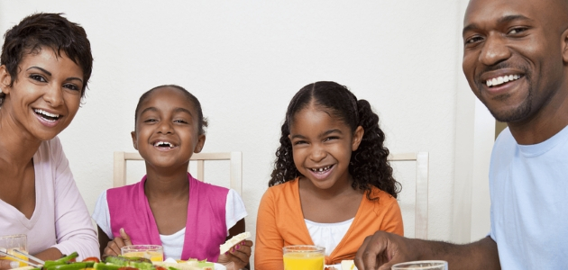 Four people sit smiling around a table, enjoying a meal together. The table is filled with food and drinks, creating a joyful, familial atmosphere.