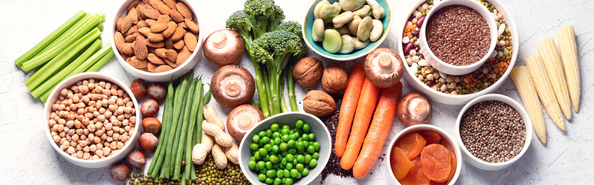 Various fresh vegetables, nuts, and legumes are arranged on a white surface, including celery, almonds, broccoli, mushrooms, carrots, peas, mixed beans, and grains in bowls.