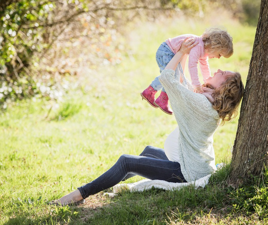 A woman sitting under a tree lifts a small child in pink boots, surrounded by a grassy field with sunlight filtering through nearby trees.