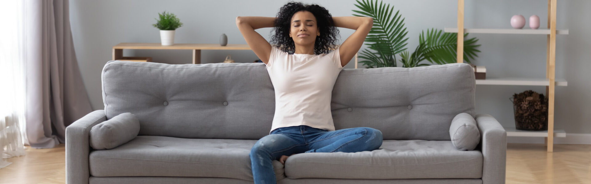 A person sits cross-legged, relaxing with arms behind head on a gray couch. The room has light curtains, green plants, and shelves with decor in the background.