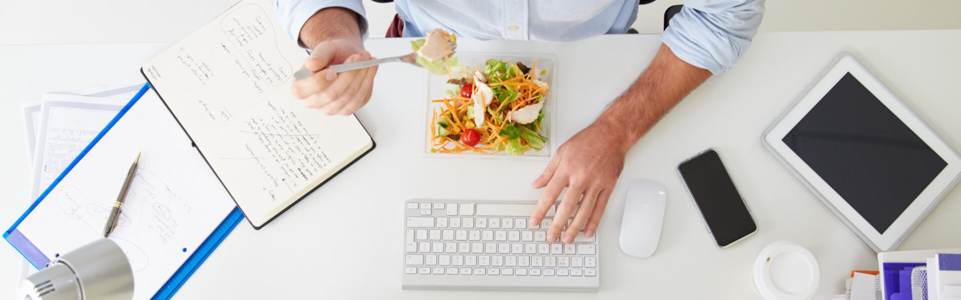 A person eats salad at a desk, using a fork with one hand while typing on a keyboard. The desk holds a notebook with notes, a tablet, a smartphone, and papers.