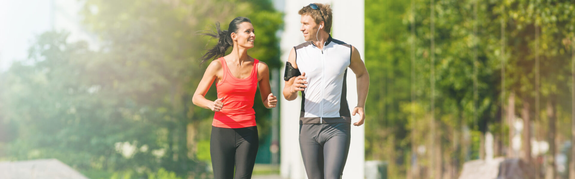 A man and a woman jog together on a sunny path surrounded by lush green trees, both smiling and wearing athletic clothing.