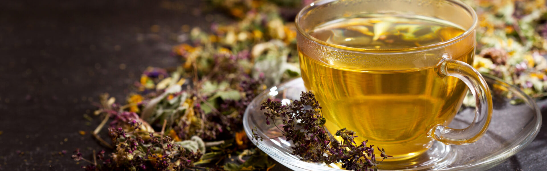 A glass cup holds steaming herbal tea, resting on a saucer surrounded by colorful, dried herbs scattered on a dark surface.