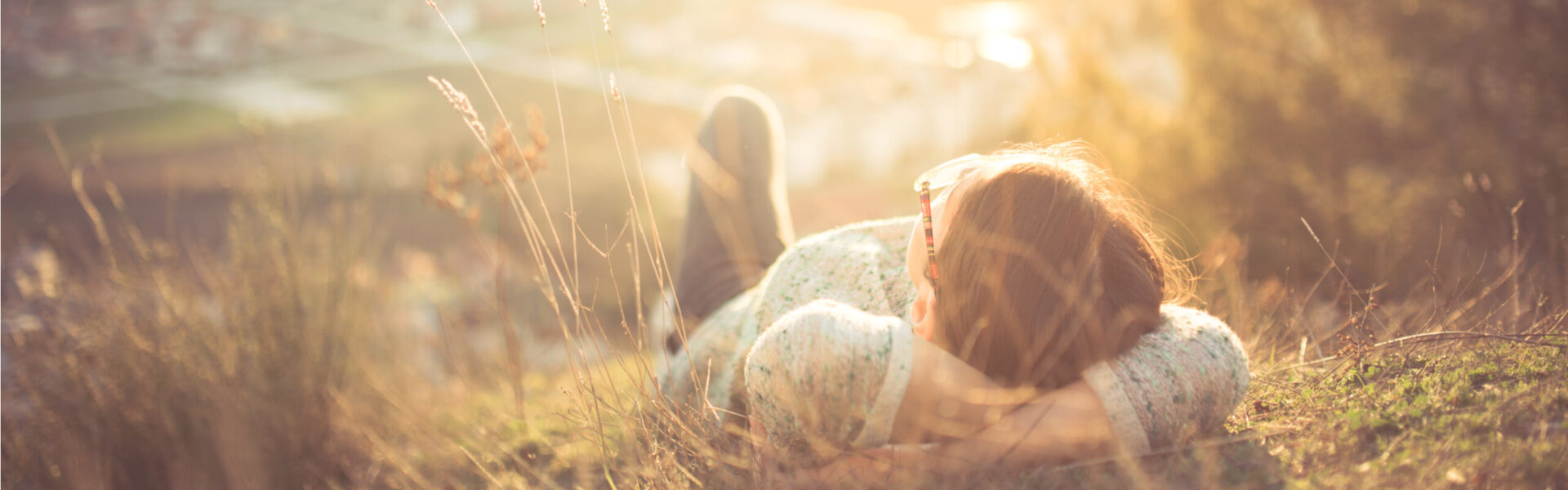 Person lying on a grassy hill, partially obscured, facing upward with hands behind head. The sun casts a warm glow, creating a peaceful, serene atmosphere.