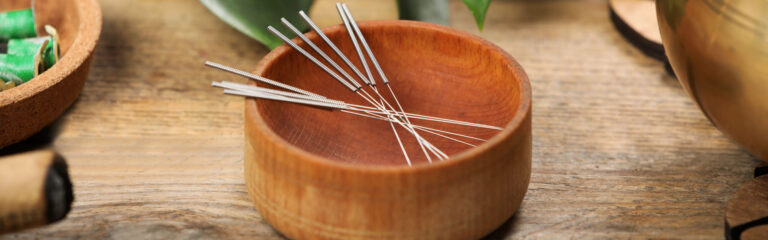 Metal acupuncture needles rest in a round wooden bowl on a wooden table, surrounded by green leaves and natural elements.