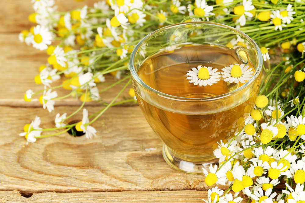 A glass cup contains chamomile tea, with two chamomile flowers floating on top, surrounded by scattered chamomile flowers on a rustic wooden surface.