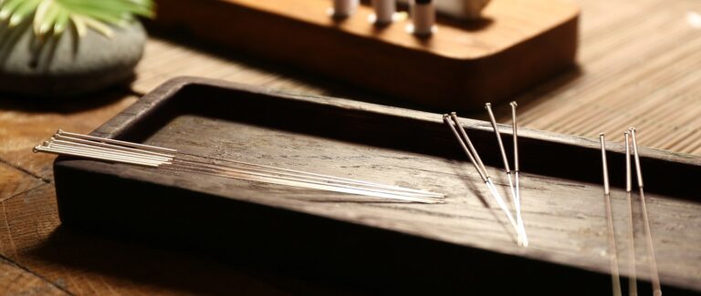 Thin acupuncture needles are neatly arranged on a wooden tray, placed on a rustic wooden table, with a blurred background featuring a small plant and additional acupuncture tools.