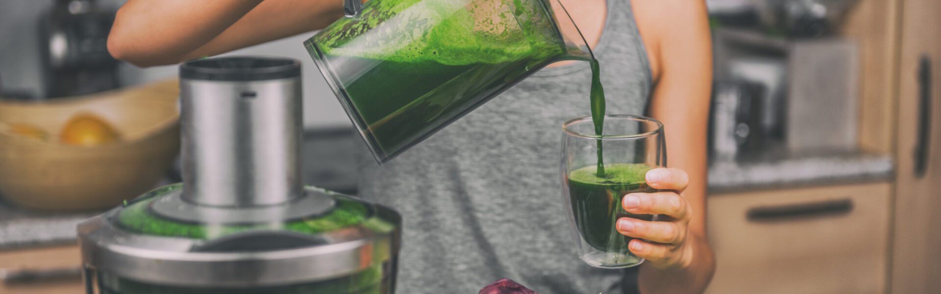 Person pouring green juice from a blender jug into a glass in a kitchen environment, with a countertop and bowl of fruit in the background.