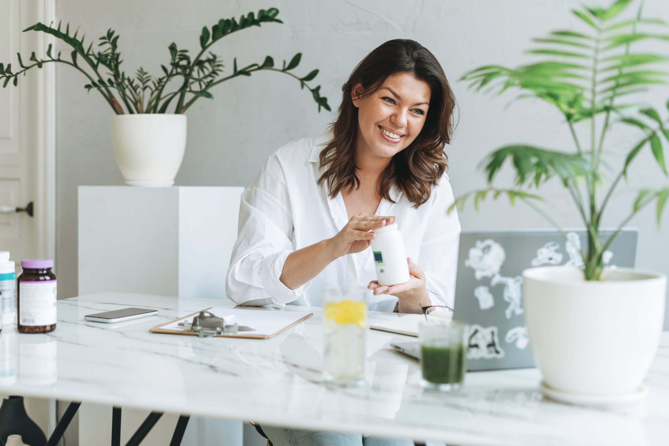 A smiling woman holds a bottle while seated at a desk with a laptop, surrounded by potted plants and health supplements in a bright, modern office setting.