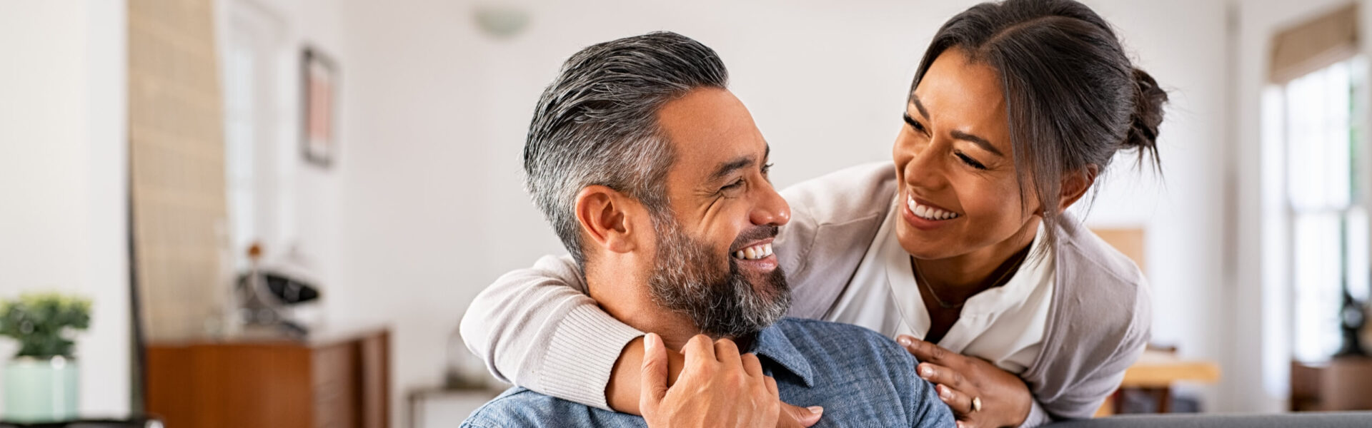 A smiling couple embraces, looking at each other joyfully in a bright, cozy living room with wooden furniture and a potted plant in the background.