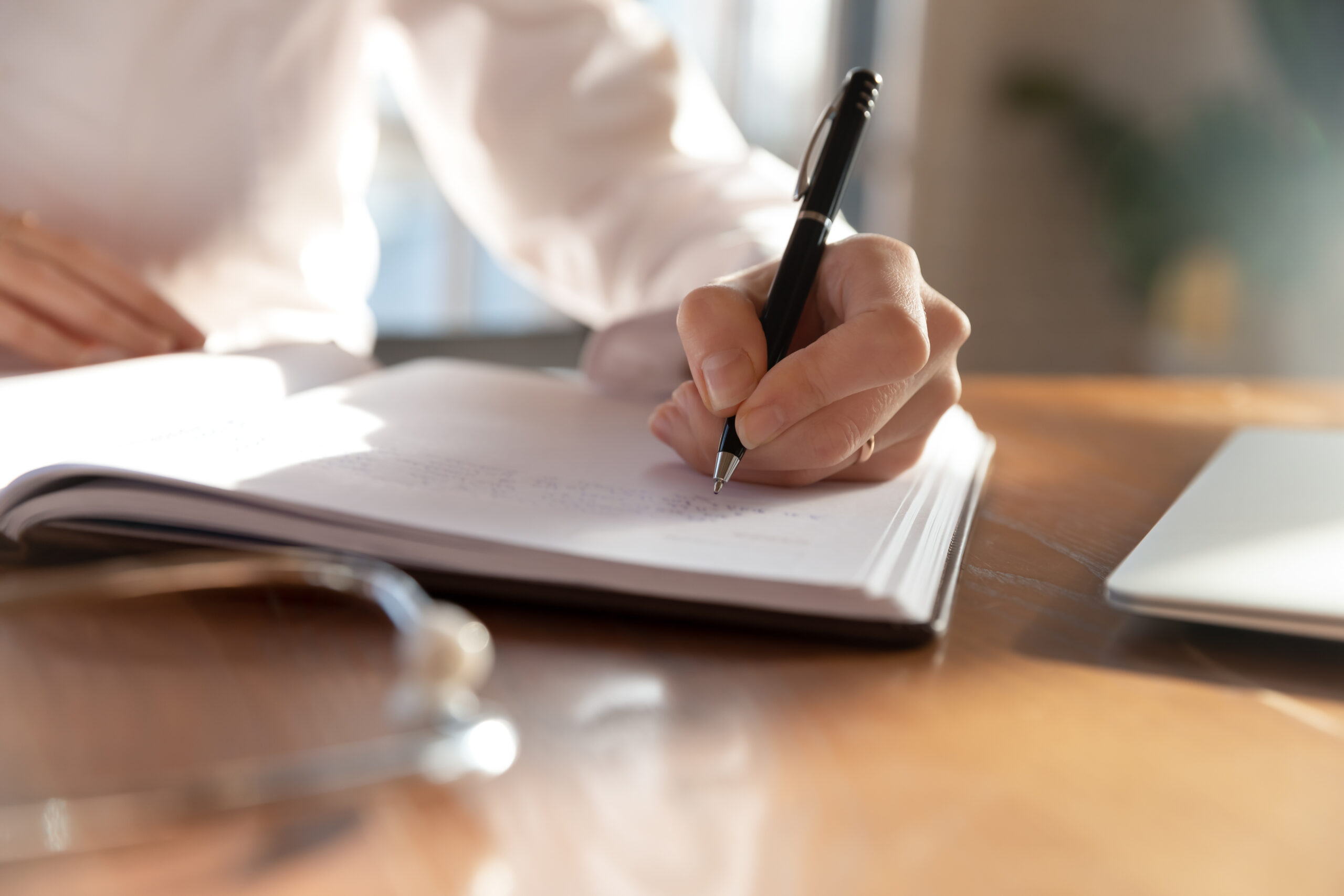 A hand writes with a pen on a notebook, resting on a wooden table with sunlight streaming through a window. An open laptop and a pair of glasses are nearby.