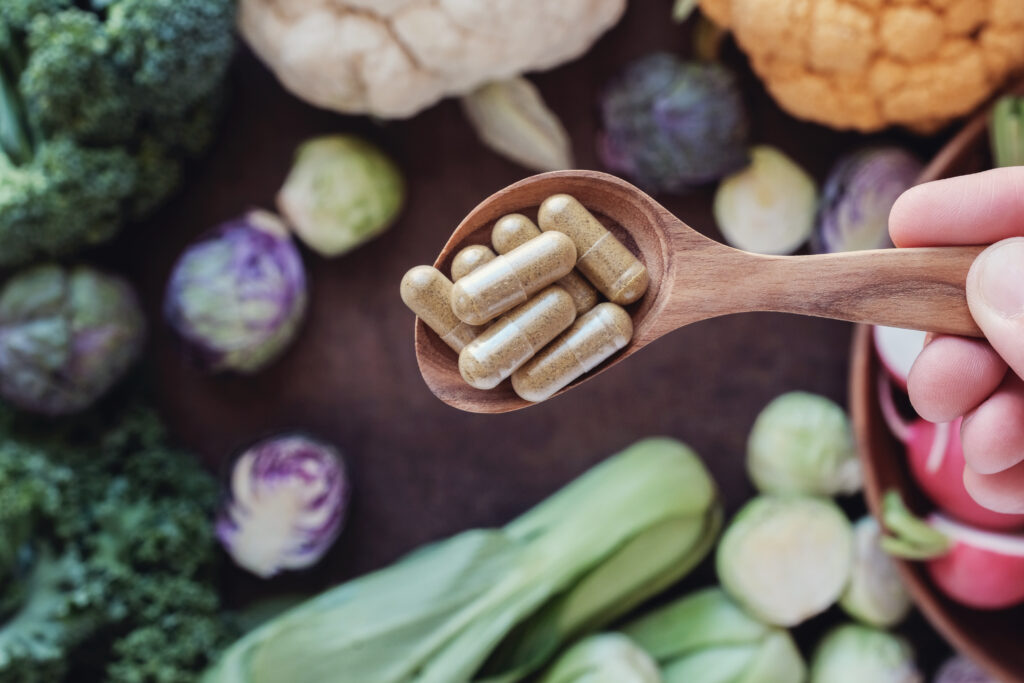 Wooden spoon holds capsules above an assortment of fresh vegetables, including broccoli, cauliflower, and bok choy, on a dark background.