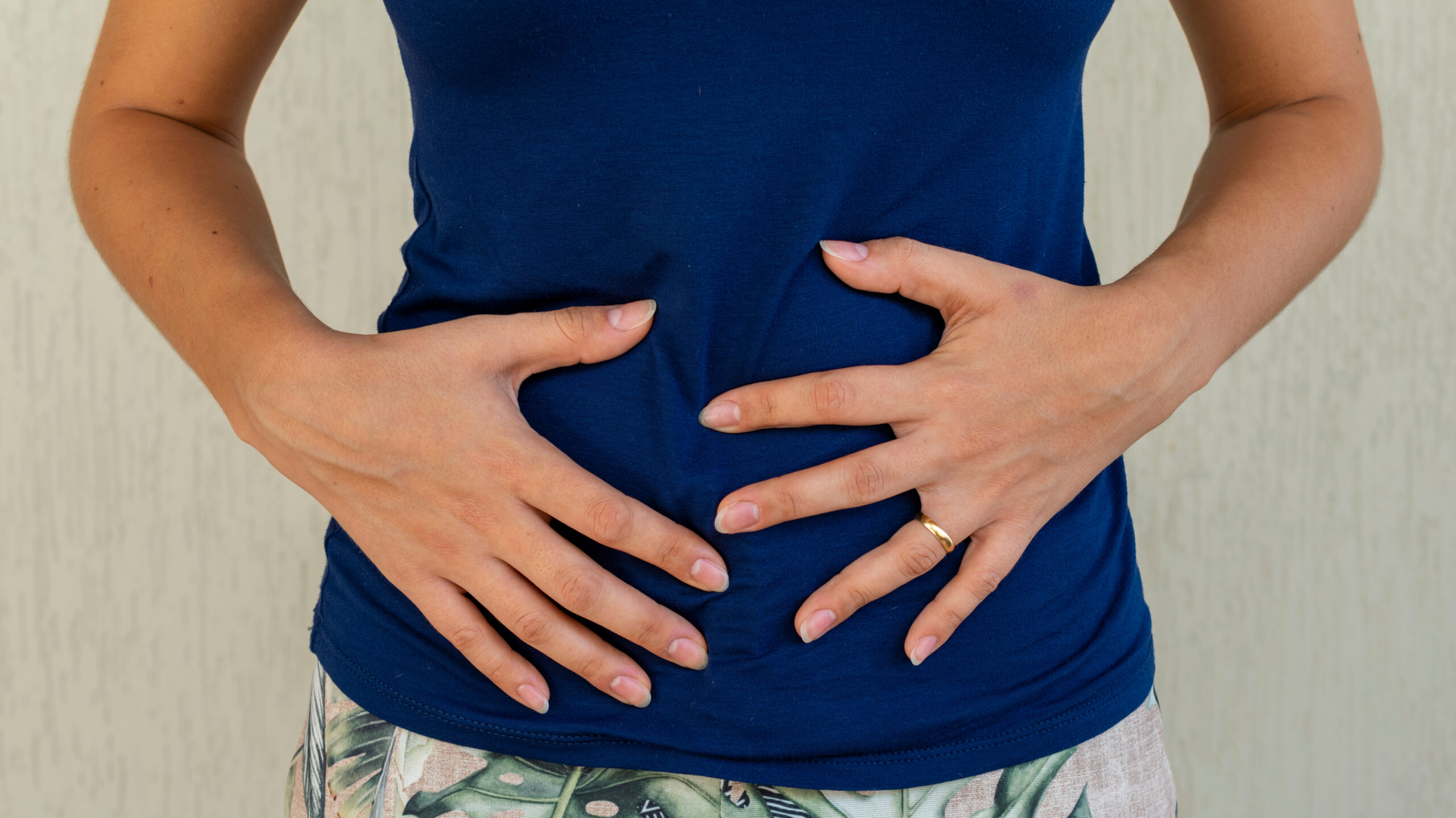 Hands resting on a person's stomach covered by a blue shirt, positioned indoors with a plain background. A gold ring is on one finger.