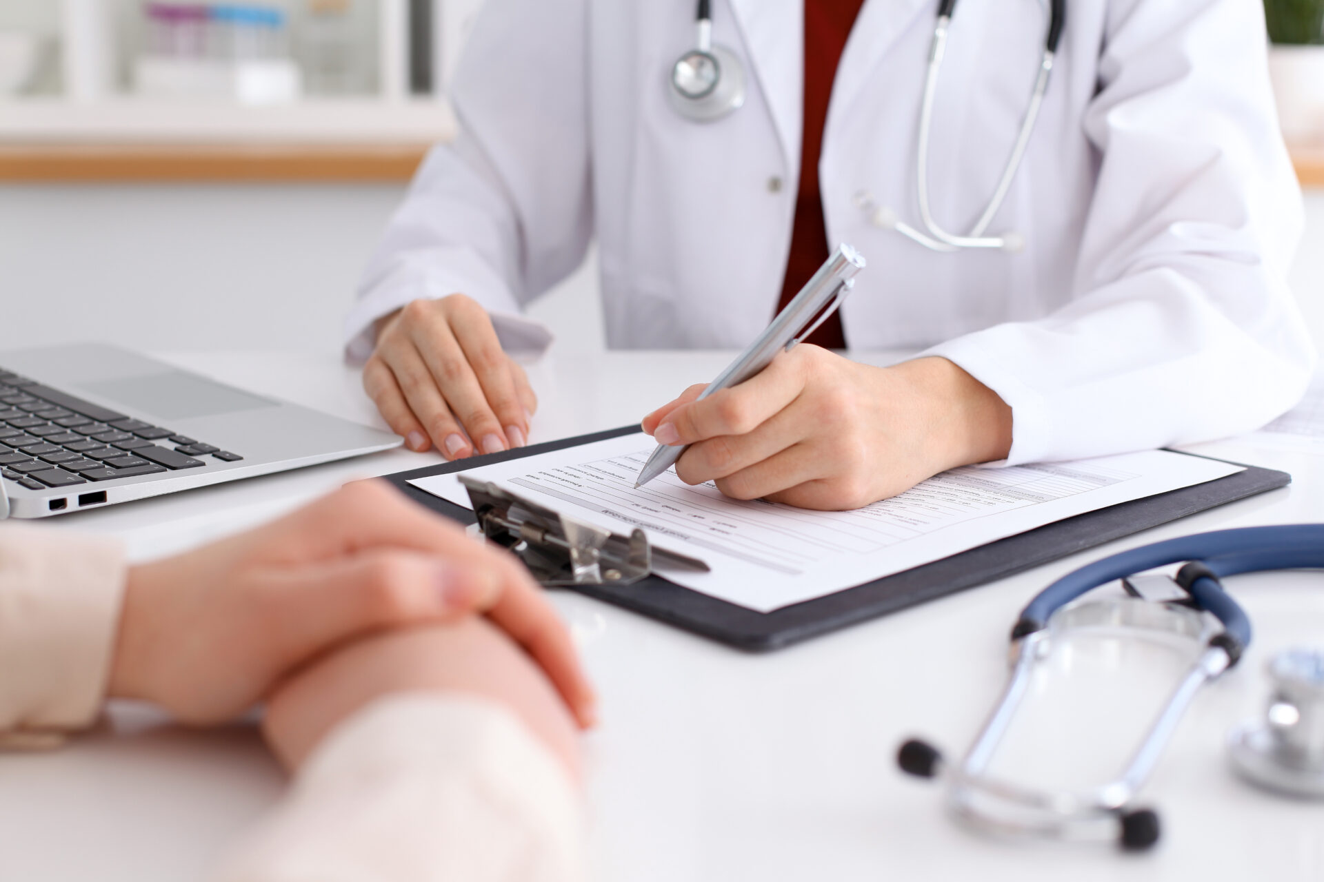 Doctor writing on a clipboard during a consultation; patient sits across the table, surrounded by a laptop and stethoscope in a clinical setting.