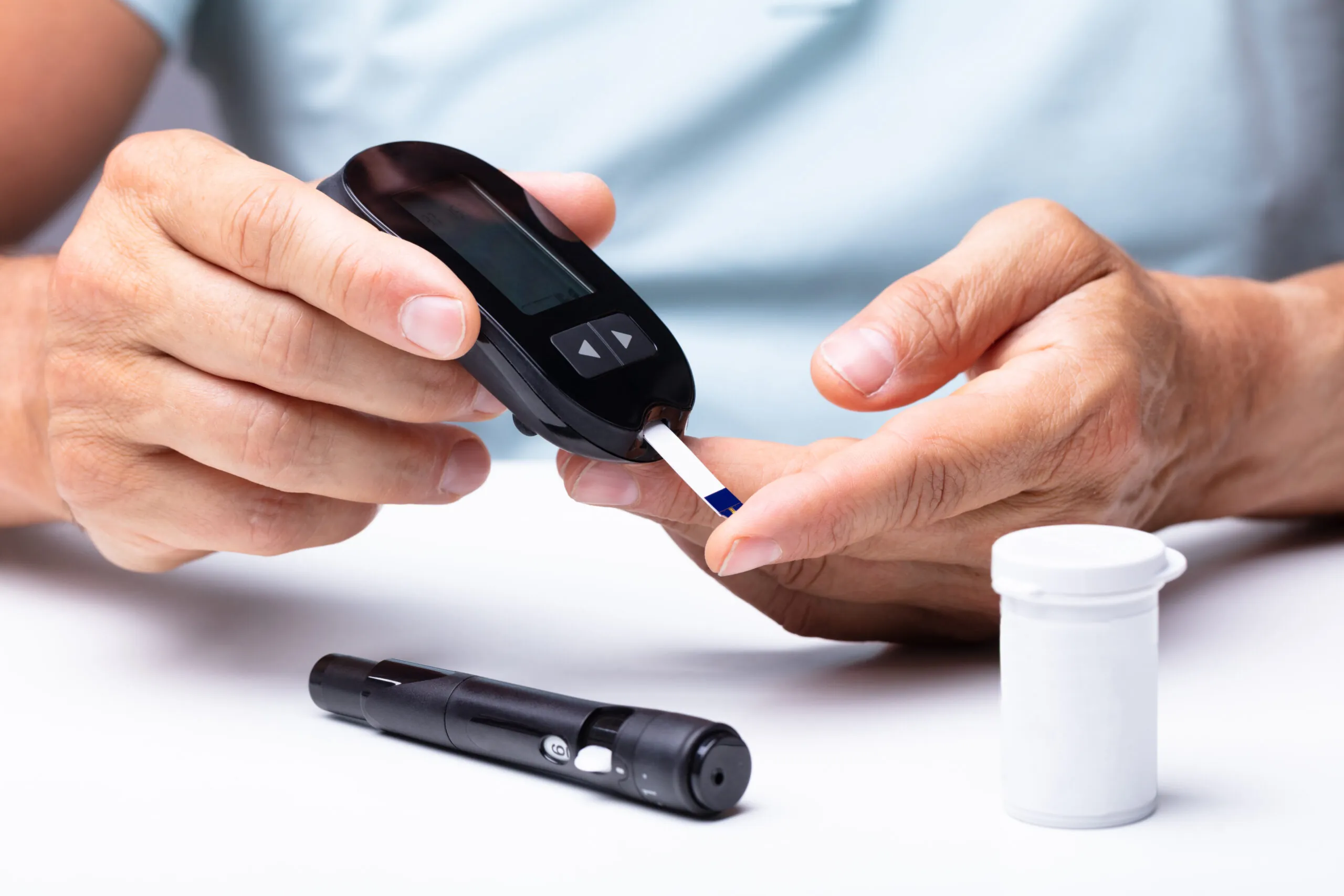 Hands hold a blood glucose meter and test strip, checking a fingertip; a white container and lancet device rest on a white table.