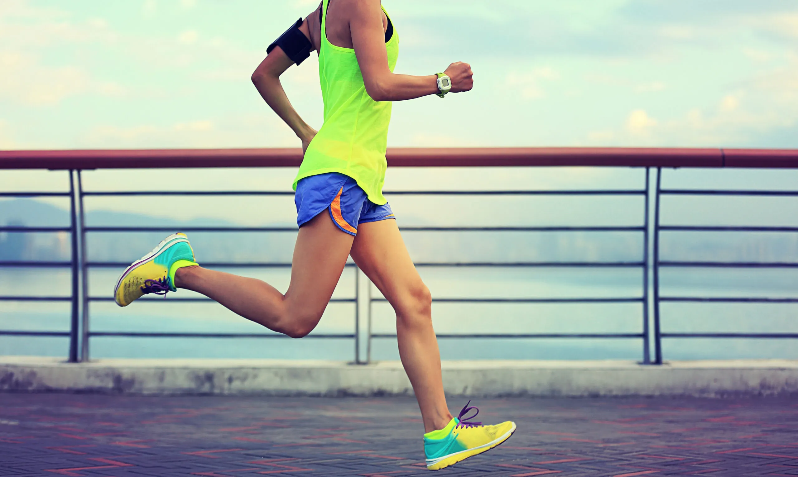 Runner wearing a bright yellow tank top and blue shorts jogs along a waterfront promenade, with a railing and distant mountains visible in the background.