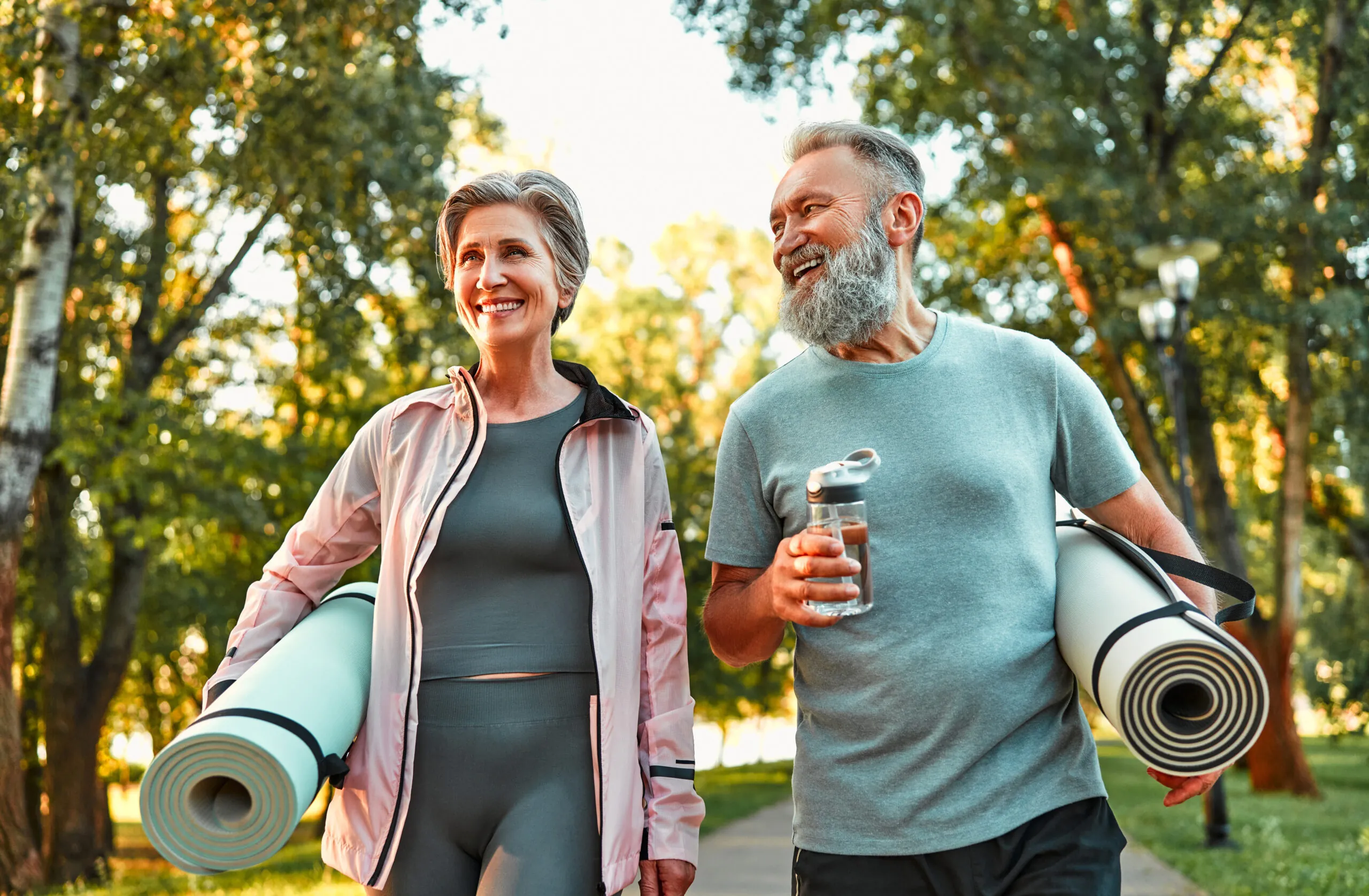 Two people carry yoga mats and walk through a sunlit park, smiling and engaged in conversation, surrounded by tall trees. The man holds a water bottle.