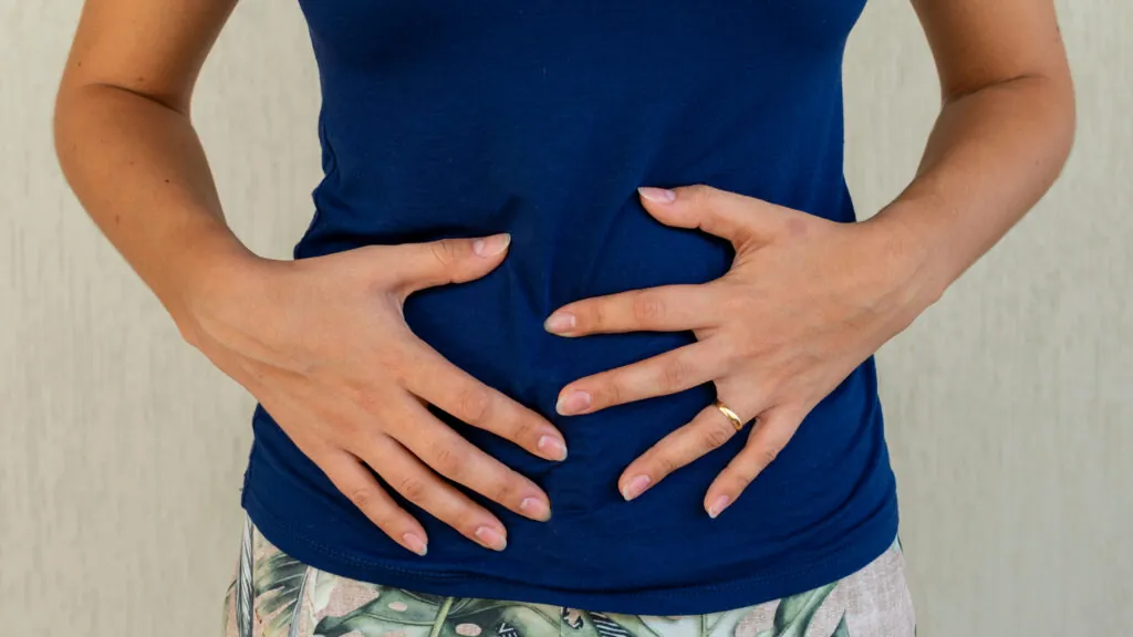 Hands resting on a person's stomach covered by a blue shirt, positioned indoors with a plain background. A gold ring is on one finger.