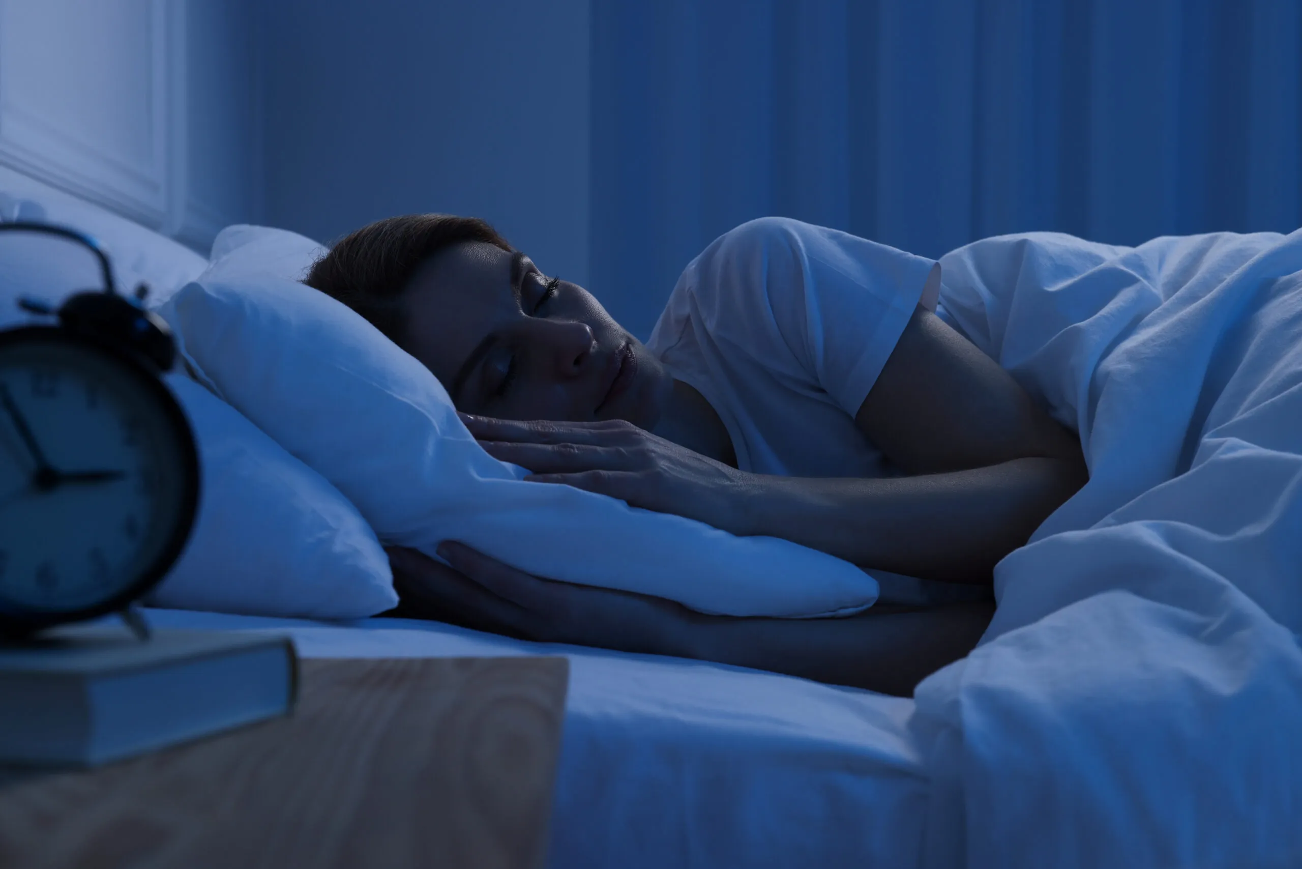 Person sleeping peacefully on a bed in dim blue lighting, with an alarm clock and book on a bedside table nearby.